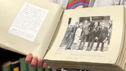 An open book shows a black and white photo of four men and a woman, one man in the centre holds up his fist in victory.