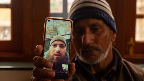  father of Azad Yousuf Kumar, holds a smart phone displaying the photos of Azad Yousuf Kumar, wearing military uniform, at Pulwama village