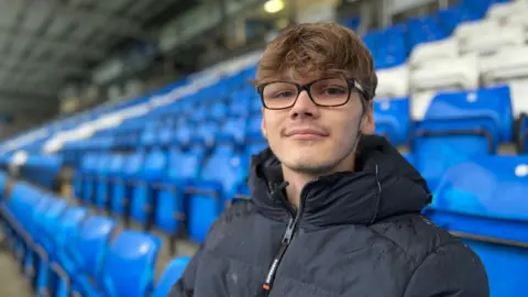 Emma Baugh/BBC 21 Year old man with dark hair and glasses wearing an anorak, sitting in the football ground seating