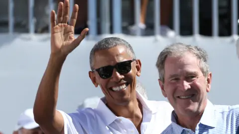 Image shows Barack Obama, who is wearing a white shirt and sunglasses, standing next to George W Bush at a golf event. Bush is wearing a blue and white striped shirt. Obama is waving to a crowd. 