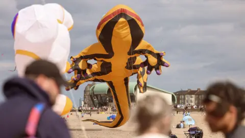 A large yellow salamander kite with purple patches on the tips of its feet. Blurred people can be seen in the foreground.