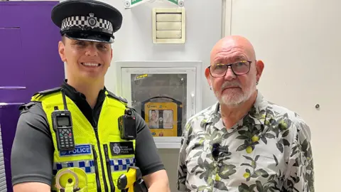 Sgt Ben Hawkins and Claude stand in front of a defibrillator at Euston station