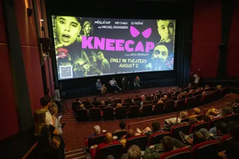 Getty Images Film critic Carlos Aguilar, writer / director Rich Peppiatt, and actors Móglaí Bap and DJ Provái attend the Film Independent Special Screening of "Kneecap" at AMC Century City 15 on July 22, 2024 in Los Angeles, California.

The group are sat at the front of a cinema, with a large Kneecap film poster behind them, and audience full of spectators