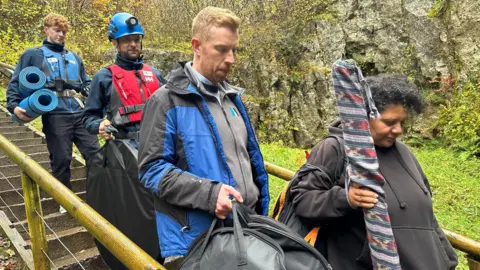 A group of people walk down an outdoor staircase. They are in a woodland, and they are carrying instruments. one person's wearing a hard hat with a torch another is carrying yoga mats.