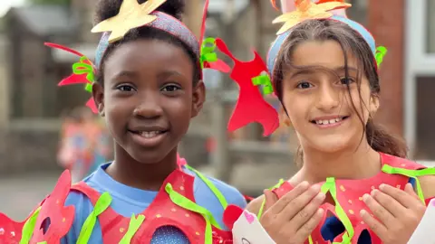 Two schoolgirls are smiling at the camera, wearing red outfits.