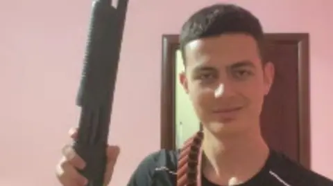 National Crime Agency Henri Nikolli holding up a shotgun while smirking. Bullets are hung around his neck and he is looking directly into the camera.