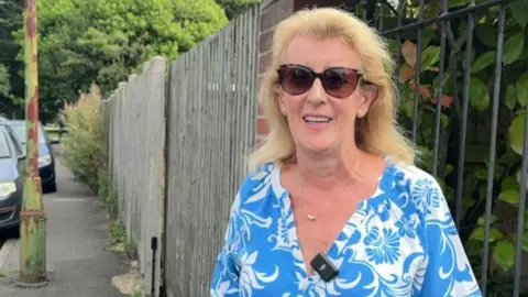 Anita Simpson standing on a pavement near two parked cars. She has long blonde hair and is wearing dark glasses and a blue and white patterned dress