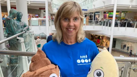 Juliette Rayner in a shopping centre, wearing a bright blue t-shirt with the ERIC logo on the front. She holds the charity's mascots, soft toys Poo and Wee in each hand, and is smiling at the camera. 