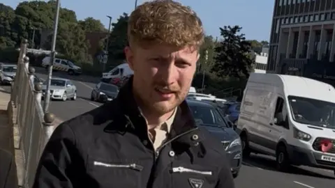 Matthew Stewart leaves court in Carlisle, outside Carlisle Magistrate's Court. He has blonde/strawberry blonde coloured short curly hair and stubble. He is wearing a black biker jacket which is large silver zips. 
