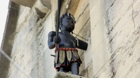 The Blackboy statue on an old church building in Stroud. It is a black statue depicting a young boy wearing a skirt made of feathers and carrying a pole to ring the bell with. He has a wire around his neck attaching him to the wall.