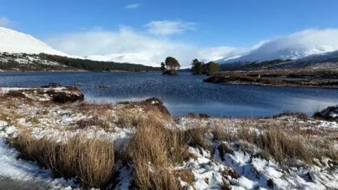 Snowy landscape with a lake and small tree-covered island under a clear blue sky.