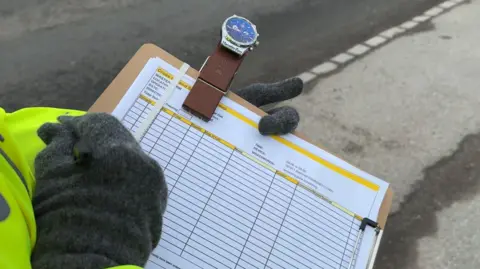 George Carden/BBC One of the volunteers wearing high vis and gloves holding a notepad with a watch clipped to it