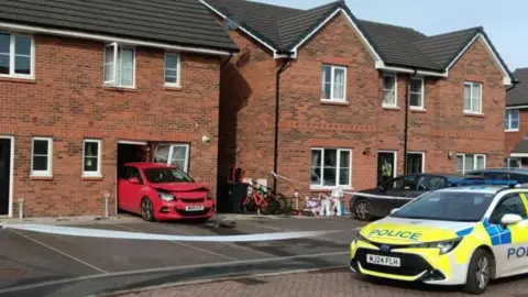 A row of semi-detached brick houses with car-parking spaces in front of them. A red car faces forward, sticking half in and half out of the front of one houses. The window to the side of it is damaged. A police car sits n the road in front of the house and there is police tape around the front of the house.