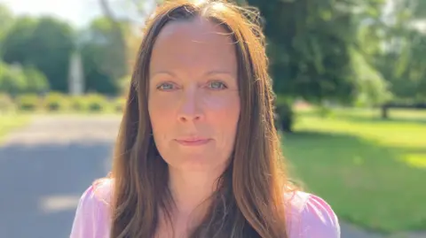 A woman with brown hair and wearing a pink top stands in a park looking down the barrel of the camera