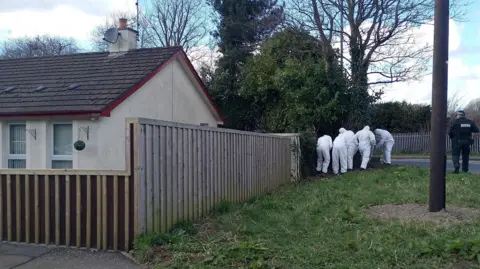 Five forensic investigators dressed white protective suits search a corner of a piece of open ground beside the house on Stream Street, Downpatrick.  A uniformed police officer stands guard beside them. 