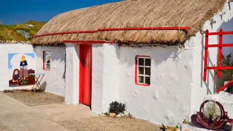 A thatched cottage with a red door and a window either side of the door. The cottage is painted white. 