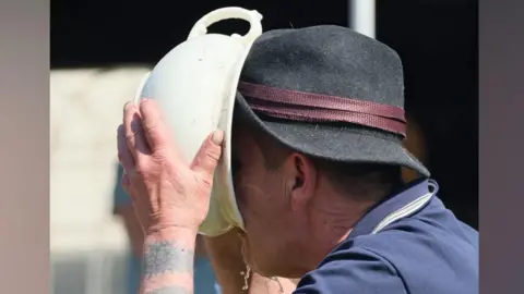 Contributed A man wearing a black hat and blue shirt drinks out of a white pot. There is some liquid dripping out of the bottom.