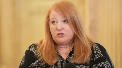 Naomi Long speaking into a microphone. She has long ginger hair and is wearing a black and white blouse and silver necklace.