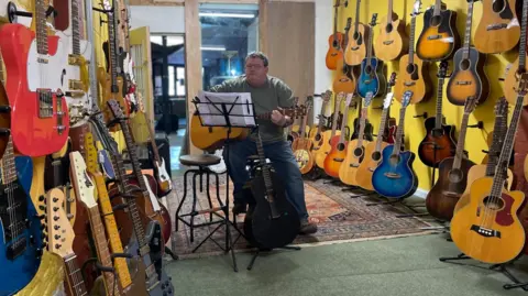 Simon Thake A heavy set man sits holding a guitar ready to play in a music shop. Acoustic and electric guitars are lined up on both sides of the shop