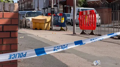 Police tape cordons off a pavement beside a pedestrian crossing in a residential street, with safety barriers, parked vehicles and rows of terraced houses visible in the background.