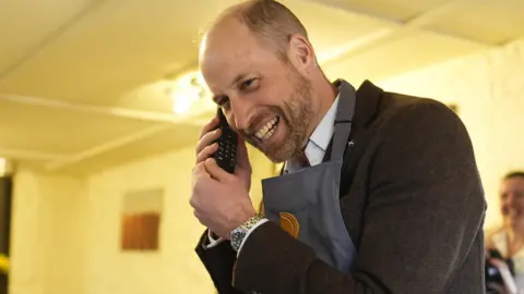 PA Media Prince William is wearing an apron and a brown jacket and is smiling while on the phone.