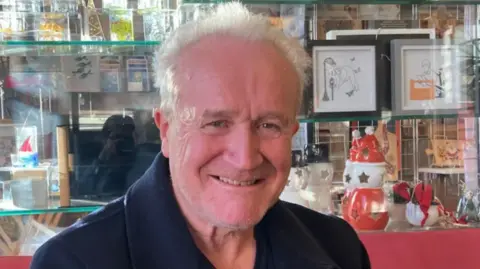 BBC/Fiona Callow A man smiling at the camera, wearing a navy blue jacket. Behind him on shelves are various trinkets and pictures. There is a mirrored wall behind the shelves.