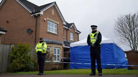 PA Media Two police officers, wrapped up against the cold, standing outside a detached, two-storey modern house. A square blue tent with a white roof is pitched on the front lawn of the house and there is police tape preventing people from entering the garden. 