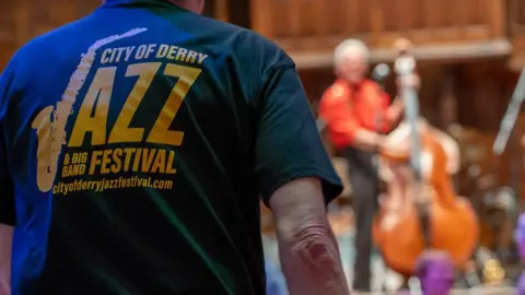 Derry City and Strabane District Council A man stands in the foreground of the image, seen from the back. He is wearing a Derry Jazz festival t shirt. In the background is a stage, where a man in a red shirt if playing a double bass