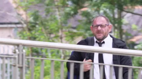 A middle -aged man with dark hair and glasses. He wears a white shirt and a black tie and holds a black backpack on one shoulder. He walks with a metal iron in front of him and green trees behind them.