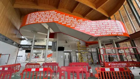 Network Rail The interior of a new railway station, which still has some scaffolding up as it is not yet complete, and red barriers on the concourse. The ceiling is wooden and beneath is a stairway balustrade, sweeping from left to right, which is coloured bright orange and has words written in white on it. 