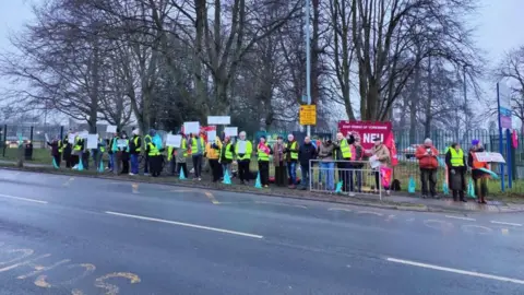 National Education Union A row of people wearing hi-vis jackets standing on the opposite side of a road on a picket line. They are standing in front of banners, and many are holding white signs.