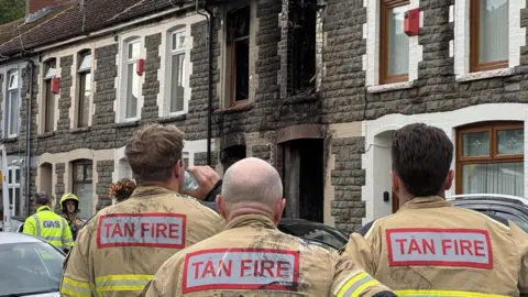 BBC A stone terraced house with fire damage around the windows and doors. Three fire officers look at the house with their backs to the camera.