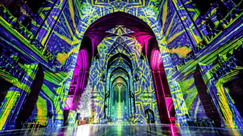 A light installation, with vivid blue and yellows colours lighting up the inside of a cathedral building. There is a central section which is lit up in pink and purple lighting.
