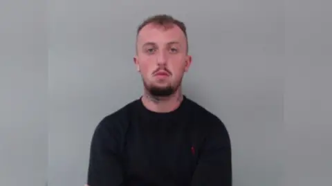 PSNI A man with short brown hair and a beard. He has tattoos on his neck and hand. He is wearing a black sweatshirt.