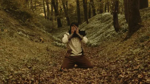 Alexander Hutchings The male character on his knees in a bed of leaves with his hands in front of his face as if in despair. He is wearing a light shirt with arm cuffs and dark trouser and is in the forest