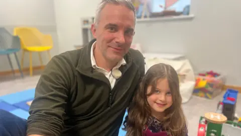 Scott, aged in his early 40s with short grey hair, wears a khaki green jumper and sits leaning in to his daughter Carey, smiling, with dark brown long hair.