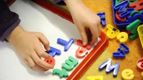 A pair of a child's hands are putting magnetic primary-coloured plastic letters on a small board. 