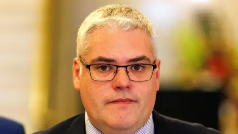 PA Media Gavin Robinson has grey hair which looks blond in the light. He is wearing dark-rimmed glasses and is unsmiling. He is wearing a suit jacket and striped shirt.