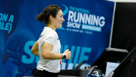 A woman wearing a white t-shirt is running on a treadmill, against a blue backdrop with signage for the National Running Show in Birmingham.