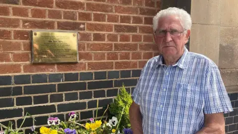 A man with short white, backcombed hair stands in front of a red brick wall with flowers beneath it. He wears a white and blue checked short-sleeved shirt. The wall has a metal plaque on it with black writing.