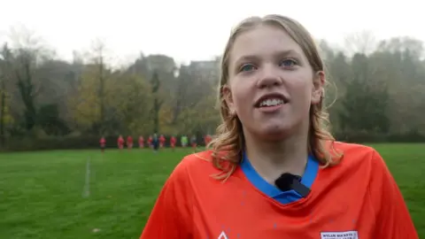 Twelve-year-old BBC Young Reporter winner Alice, wearing an orange football kit with her team in the distance behind her