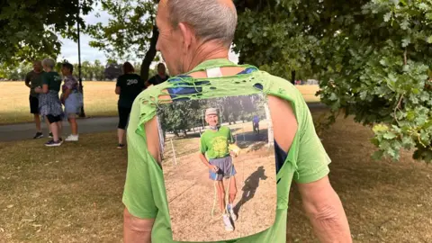 Carroll Weston/BBC A back view of Bob Emmerson who has short grey hair He is wearing a faded green T-Shirt, but most of its back is a gaping hole, while it the top is in shreds, so the black vest he is wearing can be glimpsed. The hole is largely covered by large photograph showing his father Bob Emmerson wearing the T-shirt, a cap, faded black shorts and running shoes. Behind both Bobs are trees and parched, yellow grass.