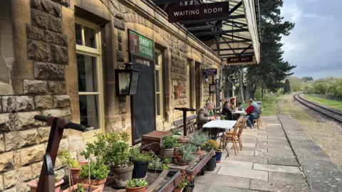 A vintage railway station on a sunny day. Some people are sitting at tables belonging to a tea shop at the end of the platform. A number of potted plants line the platform. 