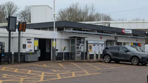 The front of Kidsgrove Railway Station, with car parking spaces in front of a single-storey white building with a grey flat roof. There are ticket machines out the front and large planters.