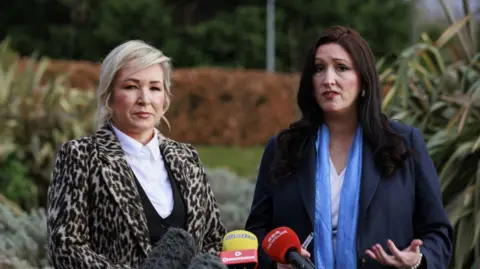 PA Media Michelle O'Neill, a woman with blonde hair, in a leopardskin jacket, with white blouse and black top, alongside Emma Little-Pengelly, a woman with dark hair, a white blouse, light blue scarf and a dark blue jacket.