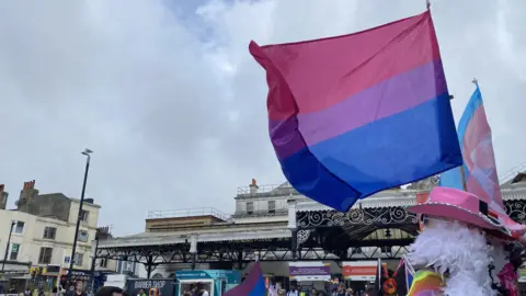 People selling rainbow flags outside Brighton Railway Station. The sky is most grey and cloudy, with a patch of blue