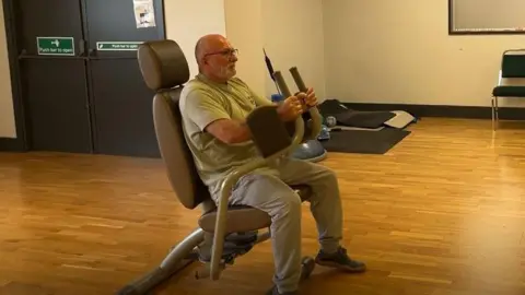 A man - Frank Carter - sat on a fitness machine in a hall. He is wearing glasses, a green shirt, light grey jogging bottoms and grey shoes. 