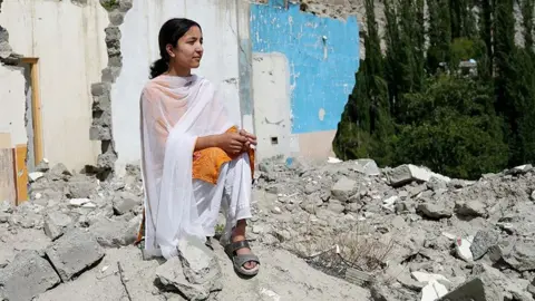 Komal Sher looks out to the right of the camera while wearing an orange top, snadals and white shawl. She is standing on the rubble of buildings in her village of Hassanabad, with damaged walls in the background.