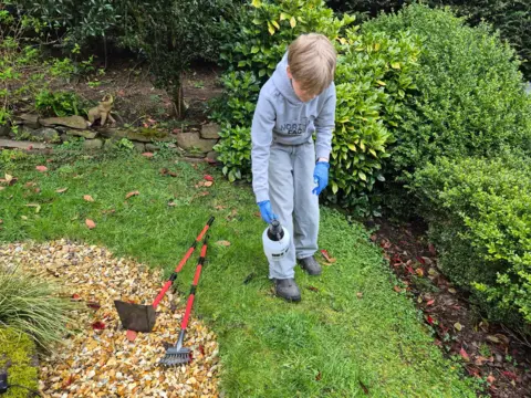 Young boy in a garden wearing a grey hoodie and blue gloves, looking down as he uses a pump sprayer on the grass, with a small rake and spade lying on a gravel patch beside him and bushes in the background