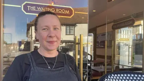 Jo Burn / BBC A woman in an apron stood in front of a glass-fronted cafe. A sign on the window of the cafe reads "THE WAITING ROOM".